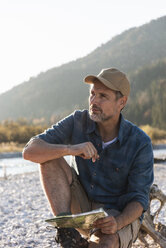 Mature man camping at riverside, sitting on tree trunk with map - UUF16291