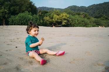 Thailand, Koh Lanta, smiling baby girl wearing UV protection shirt sitting on the beach playing with sand - GEMF02650