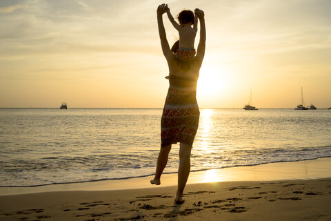 Thailand, Koh Lanta, Rückenansicht einer Mutter mit einem kleinen Mädchen auf den Schultern am Meeresufer während des Sonnenuntergangs, lizenzfreies Stockfoto