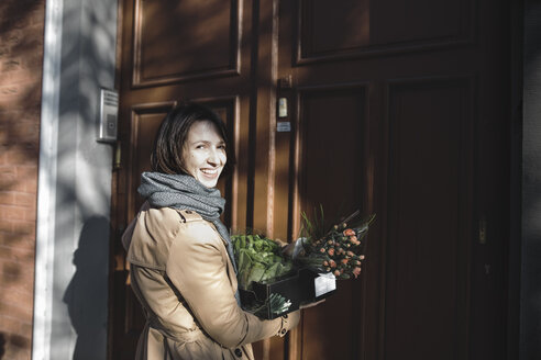 Porträt einer lachenden Frau mit einer Pappschachtel voller Einkäufe, die vor einem Hauseingang steht - KMKF00675