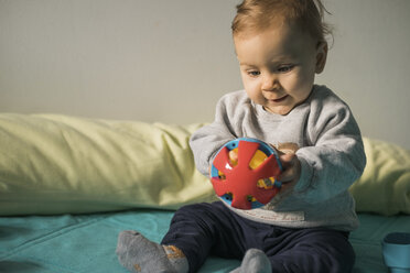 Baby-Mädchen sitzt auf dem Bett und spielt mit einem Plastikball - MOMF00559