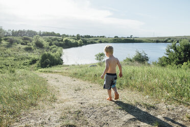 Rückansicht eines Jungen ohne Hemd, der auf einem Feld am See gegen den Himmel läuft - CAVF59901