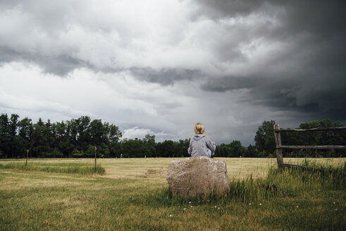 Rückansicht eines auf einem Felsen sitzenden Mädchens inmitten eines grasbewachsenen Feldes vor bewölktem Himmel - CAVF59921