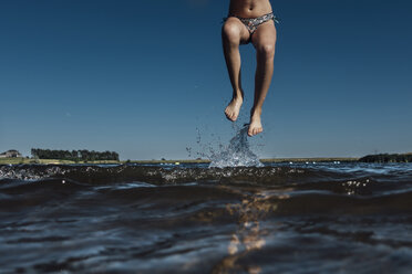 Low section of girl jumping in lake against clear blue sky - CAVF59923