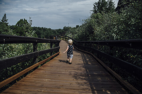 Rückansicht eines kleinen Jungen, der auf der Promenade zwischen Bäumen gegen den bewölkten Himmel läuft, lizenzfreies Stockfoto