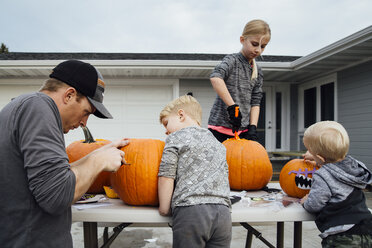 Father with kids cutting pumpkin at table in backyard during Halloween - CAVF59959