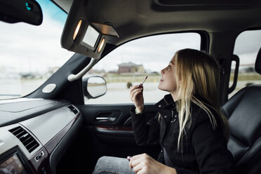 Side view of girl applying lipstick while sitting in car - CAVF59960