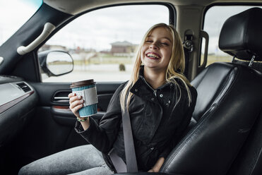 Cheerful girl holding disposable cup while sitting in car - CAVF59961