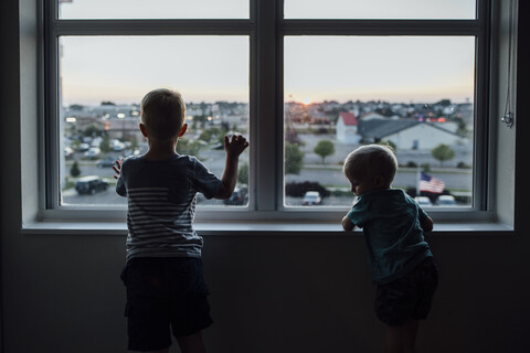 Rückansicht von Brüdern, die durch das Fenster schauen, während sie bei Sonnenuntergang zu Hause stehen, lizenzfreies Stockfoto