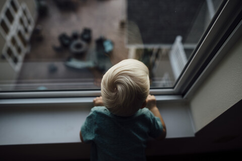 Rückansicht eines kleinen Jungen, der durch das Fenster schaut, während er zu Hause steht, lizenzfreies Stockfoto