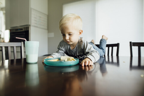 Kleiner Junge isst Essen, während er zu Hause auf dem Tisch liegt, lizenzfreies Stockfoto
