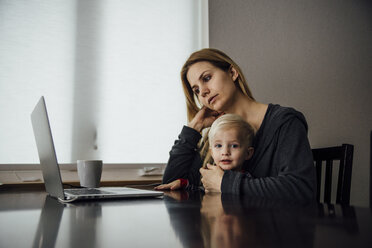 Portrait of baby boy sitting with mother using laptop on table at home - CAVF59971