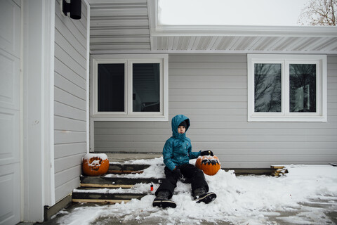 Porträt eines Mädchens in warmer Kleidung, das mit einem Halloween-Kürbis auf dem Schnee vor einem Haus im Hinterhof sitzt, lizenzfreies Stockfoto