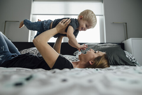 Seitenansicht einer glücklichen Mutter, die ihren Sohn abholt, während er zu Hause auf dem Bett liegt, lizenzfreies Stockfoto