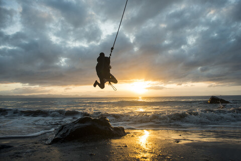 Silhouette Mann hängt an Seil über Strand gegen bewölkten Himmel bei Sonnenuntergang, lizenzfreies Stockfoto