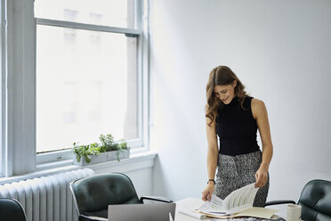 Smiling businesswoman examining documents while standing at conference table in office - CAVF60041