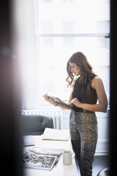 Geschäftsfrau prüft Dokumente, während sie an einem Konferenztisch im Büro steht, gesehen durch ein Fenster - CAVF60042