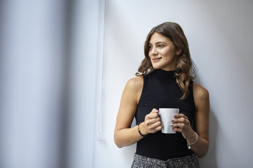 Smiling businesswoman holding coffee cup while standing against wall in office - CAVF60044
