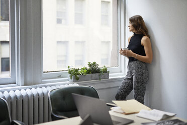 Nachdenkliche Geschäftsfrau mit Kaffeetasse, die durch das Fenster schaut, während sie an der Wand im Büro steht - CAVF60045