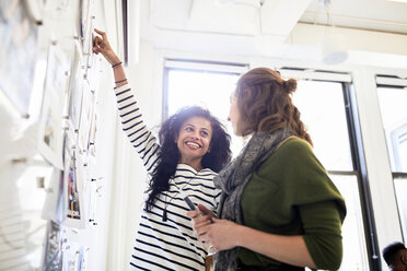 Smiling businesswoman looking at female colleague while standing by bulletin board in office - CAVF60067