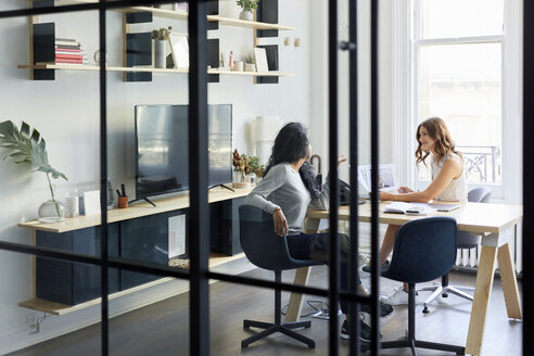 Weibliche Kollegen arbeiten zusammen, während sie am Schreibtisch im Büro sitzen, gesehen durch ein Fenster - CAVF60075