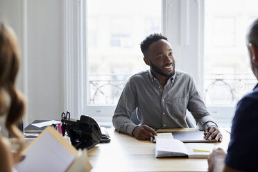 Smiling businessman talking with colleague at desk against window in office - CAVF60081