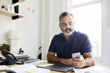 Businessman using smart phone while sitting at desk in creative office - CAVF60089