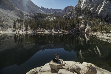 High angle view of woman with dog sitting on rock by lake during sunny day - CAVF60136