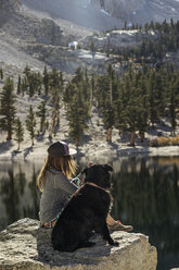 Seitenansicht einer Frau mit Hund, die auf einem Felsen am See sitzt, an einem sonnigen Tag - CAVF60137