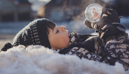 Seitenansicht eines Jungen mit Schneekugel, der im Winter im Freien liegt - CAVF60185