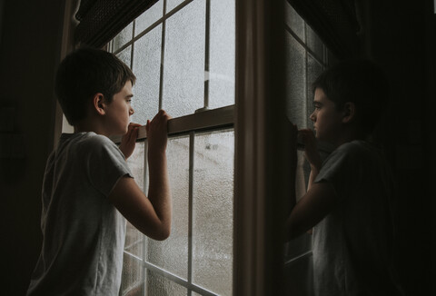 Seitenansicht eines nachdenklichen Jungen, der durch ein nasses Fenster schaut und sich im Spiegel zu Hause spiegelt, lizenzfreies Stockfoto