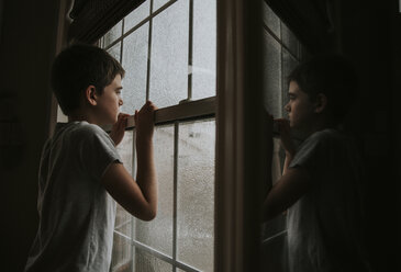 Seitenansicht eines nachdenklichen Jungen, der durch ein nasses Fenster schaut und sich im Spiegel zu Hause spiegelt - CAVF60188