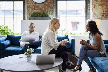 Businesswomen discussing and male colleague working on laptop while sitting at sofa in office - CAVF60212