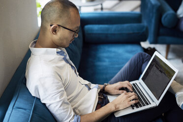 High angle view of businessman using laptop while sitting on blue sofa at creative office - CAVF60215