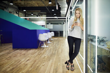 Geschäftsfrau in voller Länge, die ein Smartphone benutzt, während sie sich an eine Glaswand im Korridor lehnt - CAVF60224