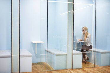 Serious businesswoman talking on mobile phone while using laptop in cubicle at office - CAVF60227