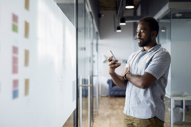 Junger Geschäftsmann, der ein Smartphone benutzt, während er im Bürokorridor steht - CAVF60243