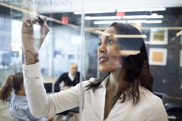 Businesswoman writing plan on glass in board room with colleagues in background - CAVF60260