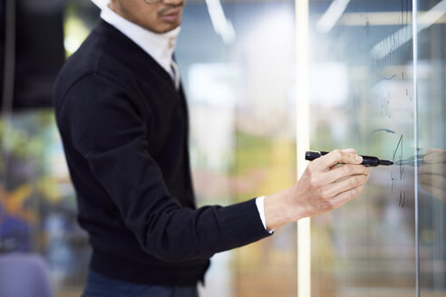 Mittelteil eines Geschäftsmannes, der bei der Arbeit im Büro auf eine Glaswand schreibt - CAVF60268