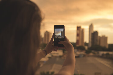 Rear view of woman photographing buildings with smart phone against sky during sunset - CAVF60309