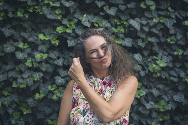 Portrait of mature woman standing in front of wall overgrown with ivy pouting mouth - JUNF01631