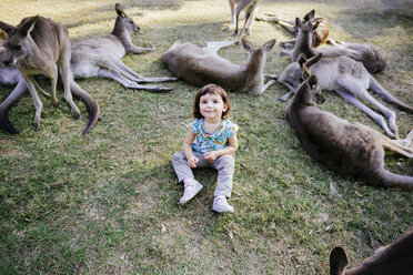 Australien, Brisbane, Porträt eines lächelnden kleinen Mädchens, das auf einer Wiese zwischen zahmen Kängurus sitzt - GEMF02684