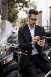 Businessman at motor scooter in the city using cell phone - MAUF01933