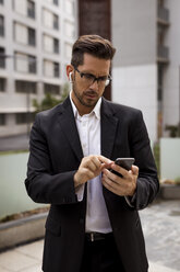 Businessman with earbuds in the city checking cell phone - MAUF01936