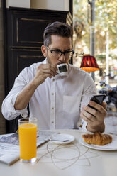 Businessman having breakfast in a cafe and checking cell phone - MAUF01955