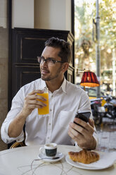 Geschäftsmann frühstückt in einem Café und hält ein Mobiltelefon in der Hand - MAUF01957