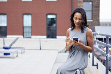 Geschäftsfrau, die ein Smartphone benutzt, während sie auf der Terrasse eines Gebäudes sitzt - CAVF60387