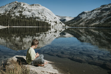 Seitenansicht eines Jungen, der auf einem Felsen am See vor den Bergen im Yosemite-Nationalpark sitzt - CAVF60426