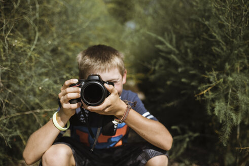 Boy photographing with camera while crouching amidst plants on field during sunny day - CAVF60448