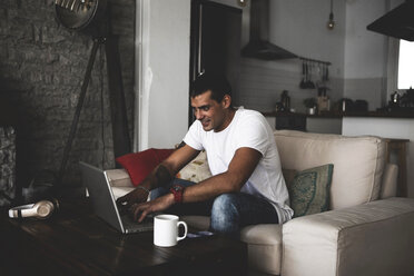 Smiling young man sitting on sofa at home using laptop - ERRF00356